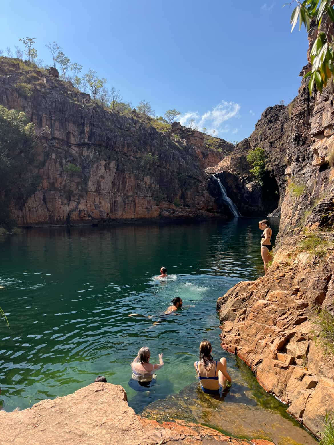 A rocky canyon with steep cliffs surrounds a clear green pool beneath a bright blue sky. A small waterfall flows into the pool, where several people are swimming and sitting on the rocks along the water’s edge.