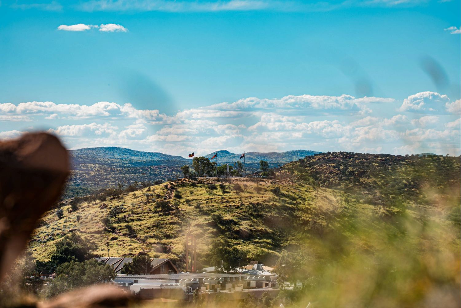View across a grassy hilltop with three flagpoles on a ridge, overlooking buildings and rolling ranges under a bright blue sky dotted with white clouds.