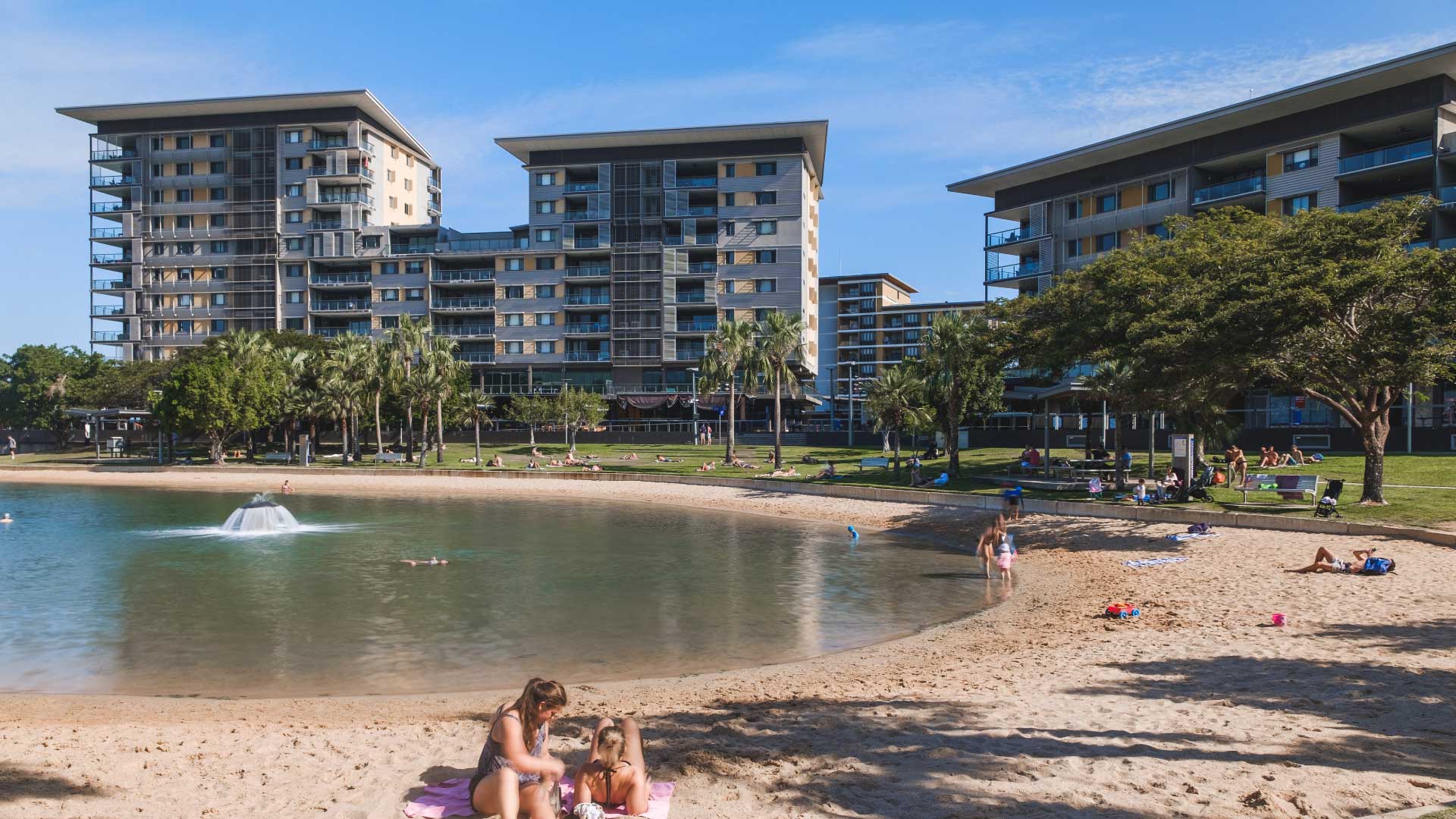 Modern waterfront apartments overlooking the sandy Darwin Waterfront beach lagoon where people swim, sunbathe and relax on the grass under palm trees on a sunny day.