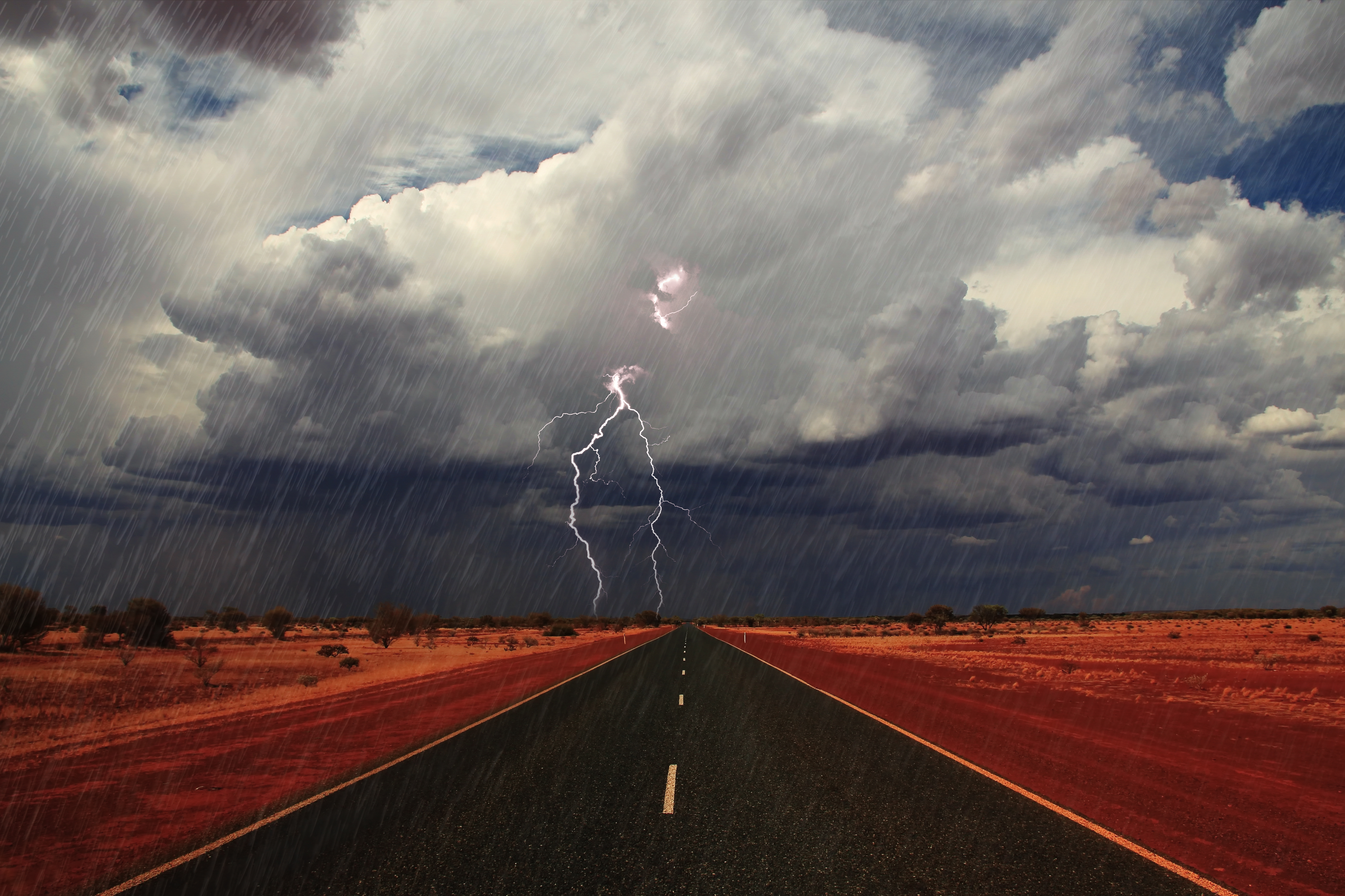 A long outback road leading to dark storm clouds as lightning strikes in the distance.