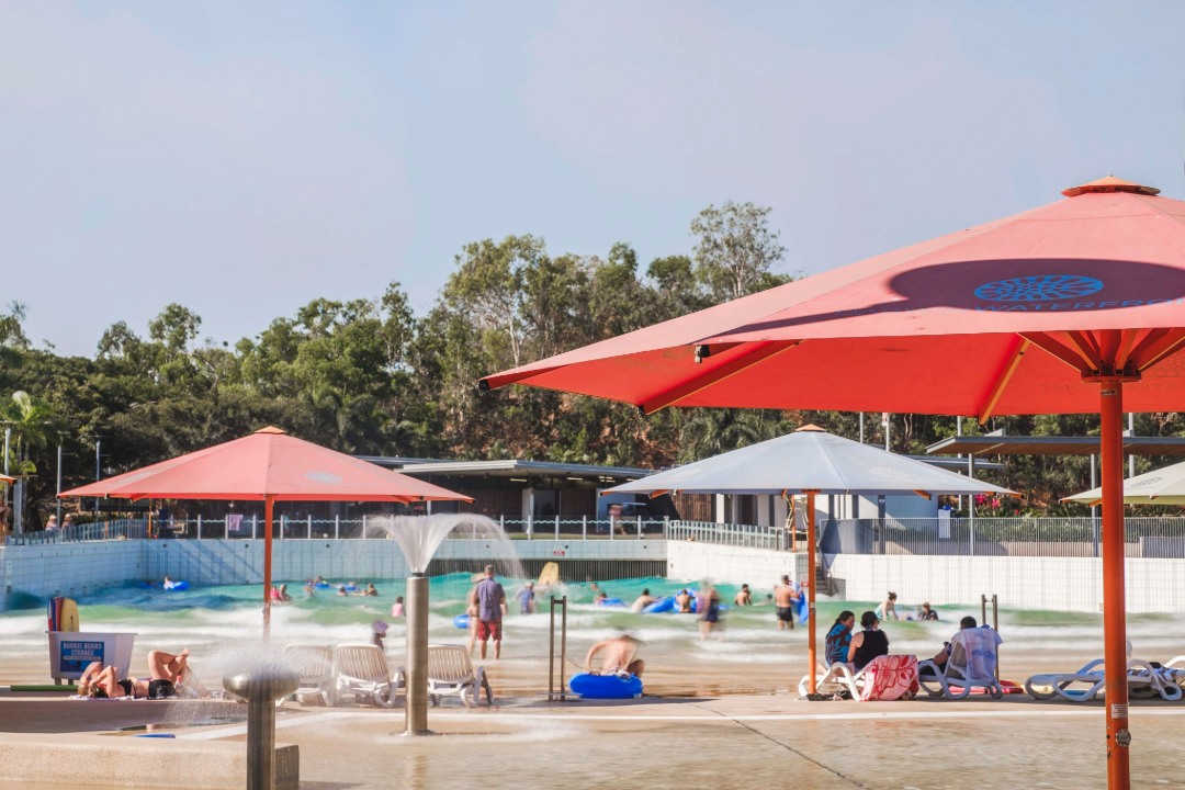A busy outdoor water park with people swimming and playing in a wave pool, surrounded by large red and white umbrellas, lounge chairs, and trees.