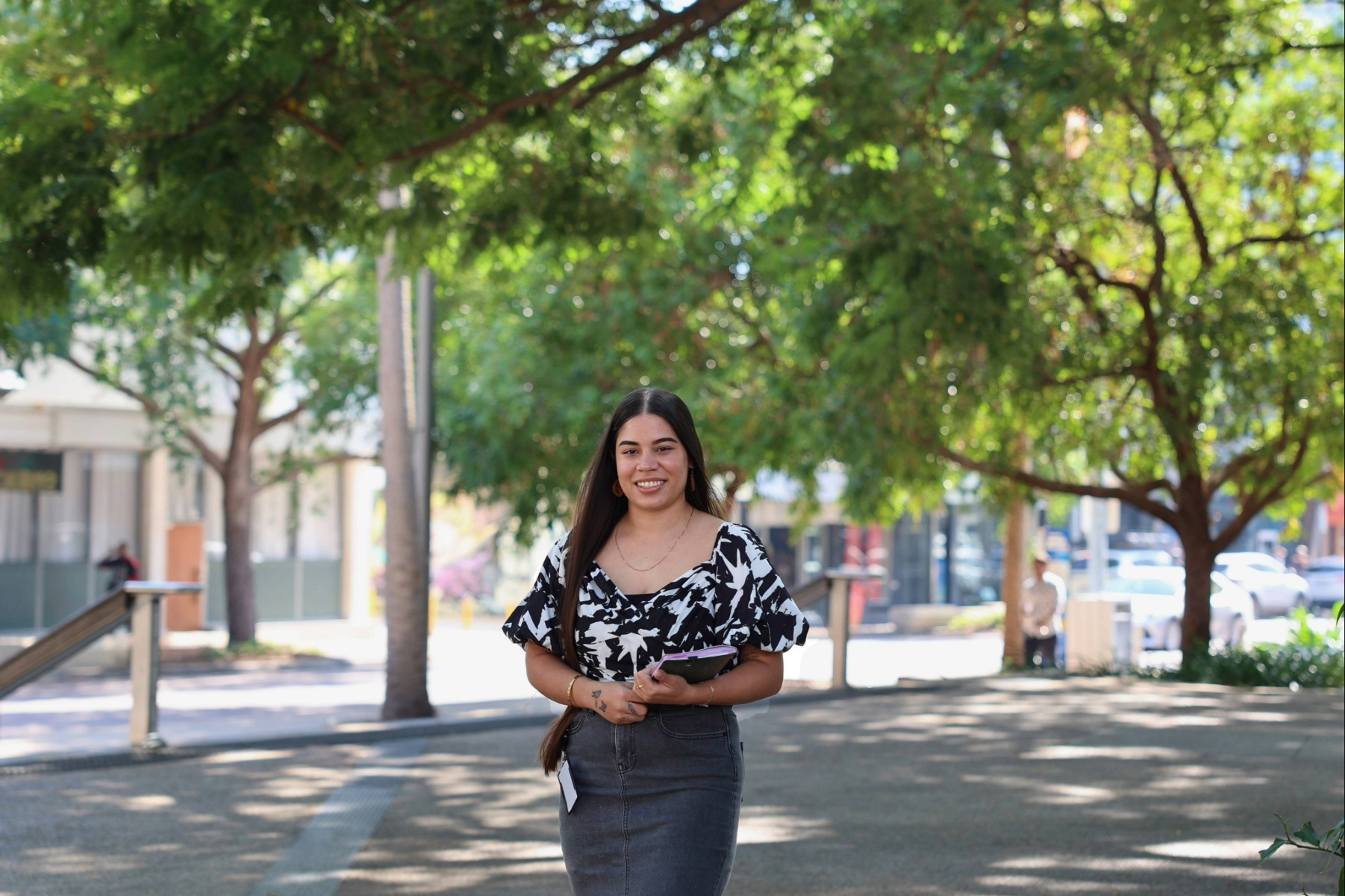 A lady standing on a shaded pedestrian pathway, holding a tablet, with trees and buildings in the background.