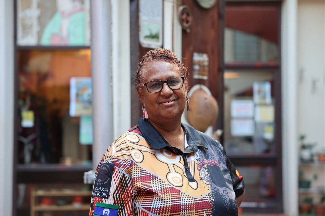 A lady, Cassandra Henaway, wearing a patterned collared shirt and glasses standing outdoors in front of a shopfront.