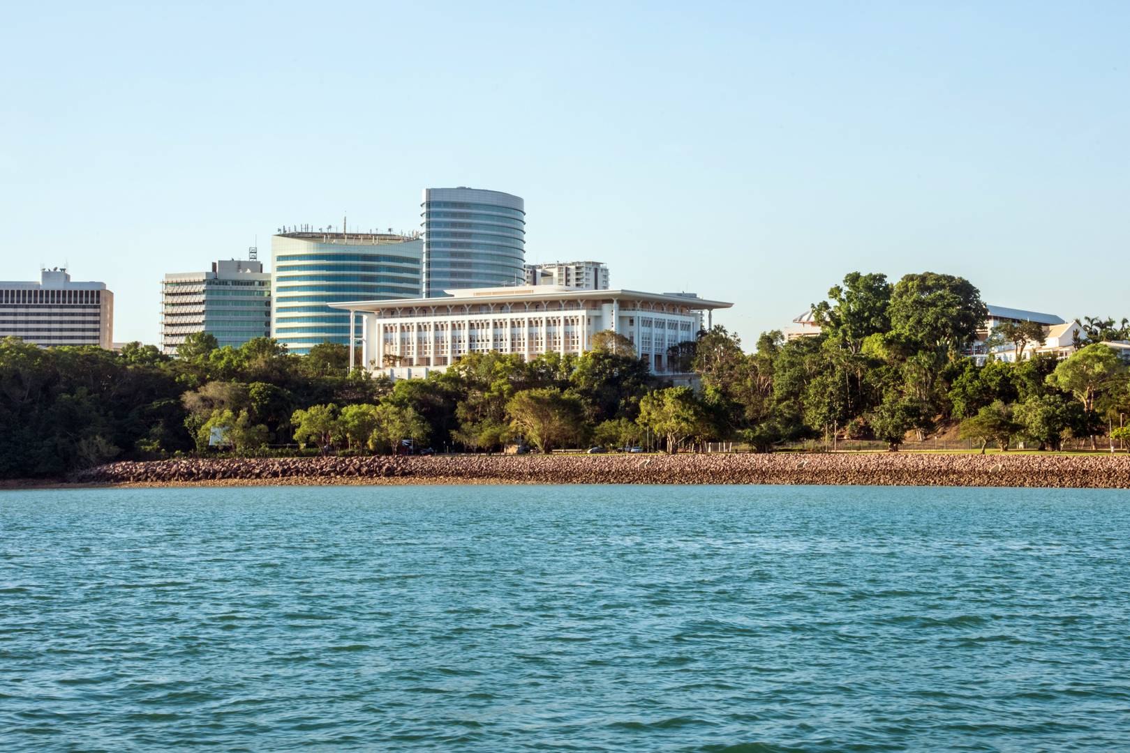 A coastal cityscape with modern buildings, trees, and calm water in the foreground.