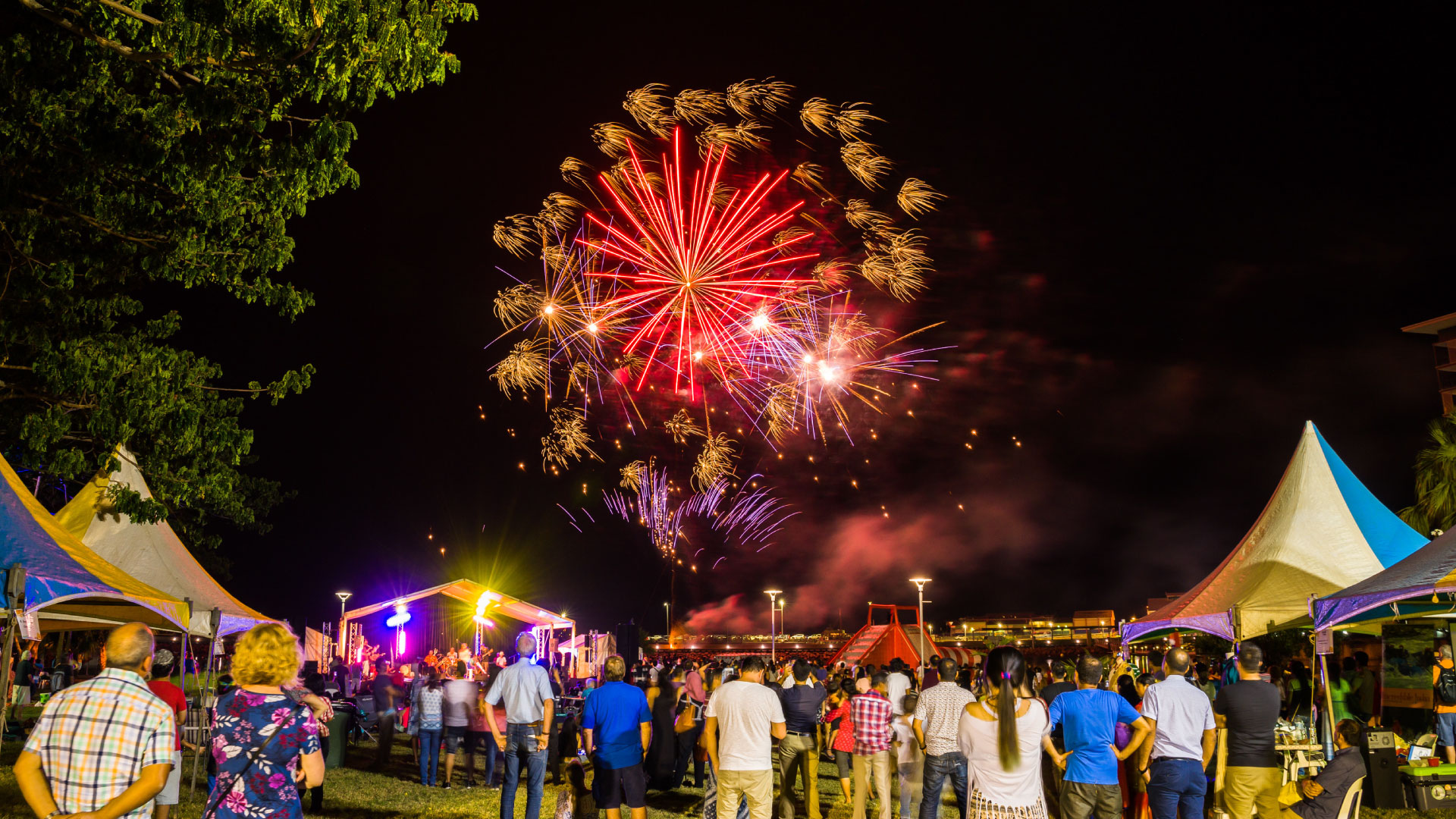 Crowd watching colorful fireworks at a lively outdoor night festival with tents, stage, and trees at the Darwin Waterfront.