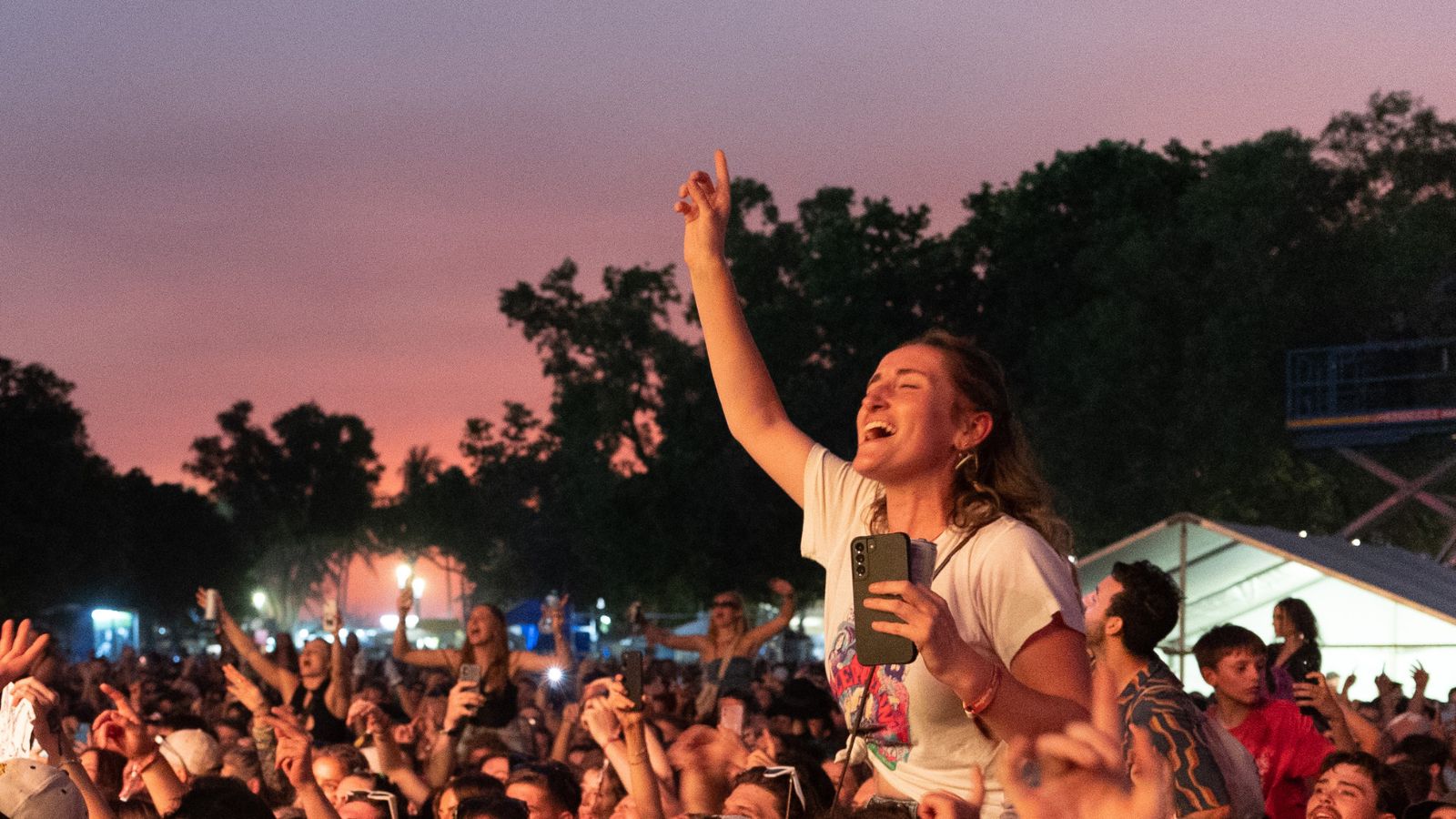 A woman sitting on someone’s shoulders cheers with her phone in hand at a crowded outdoor music event at sunset.