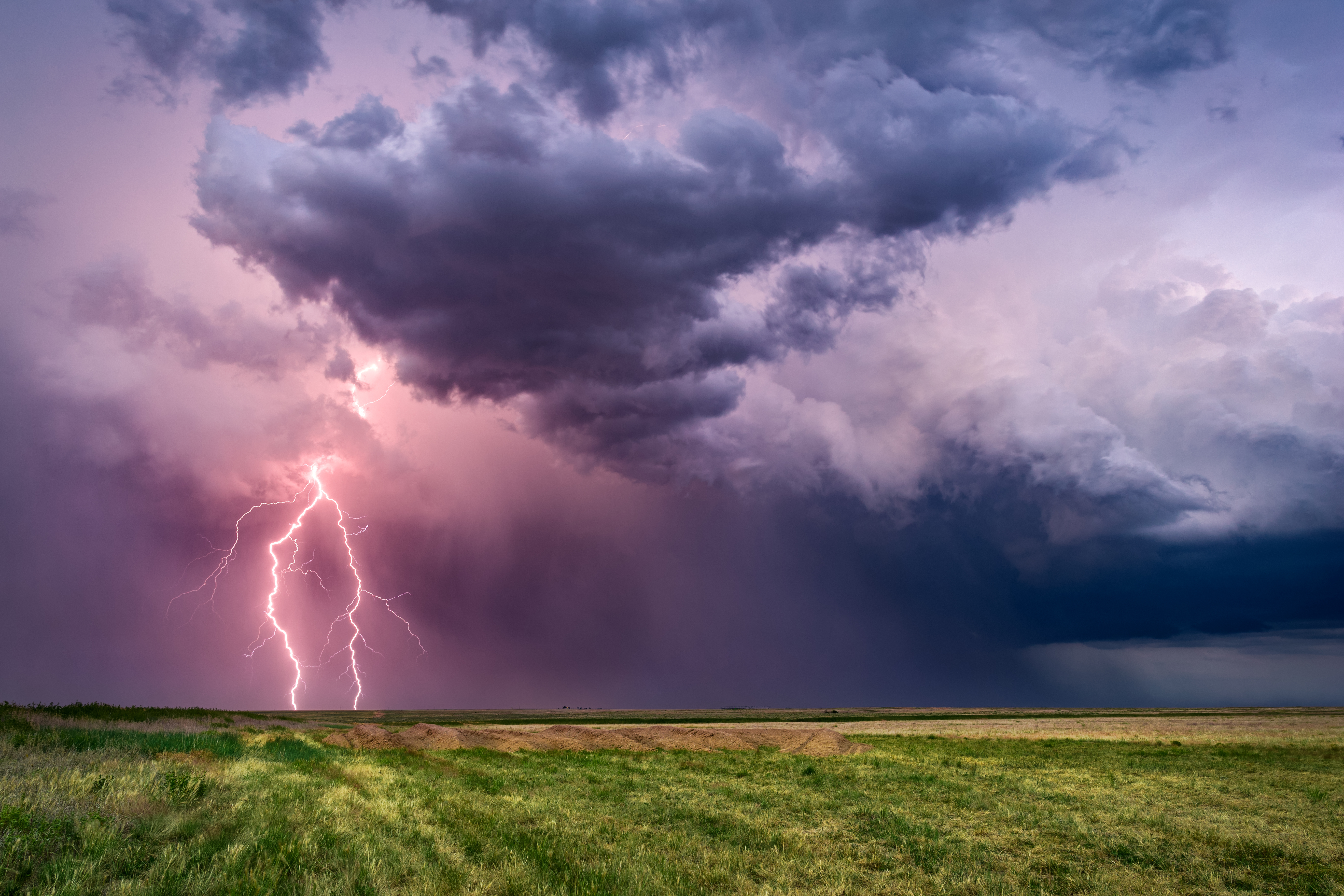 A bright lightning bolt strikes under purple and gray storm clouds over an open field.