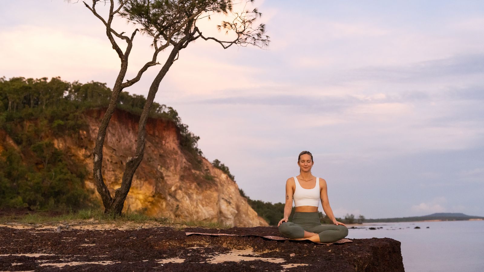 Person meditating on a rocky ledge beside coastal cliffs.