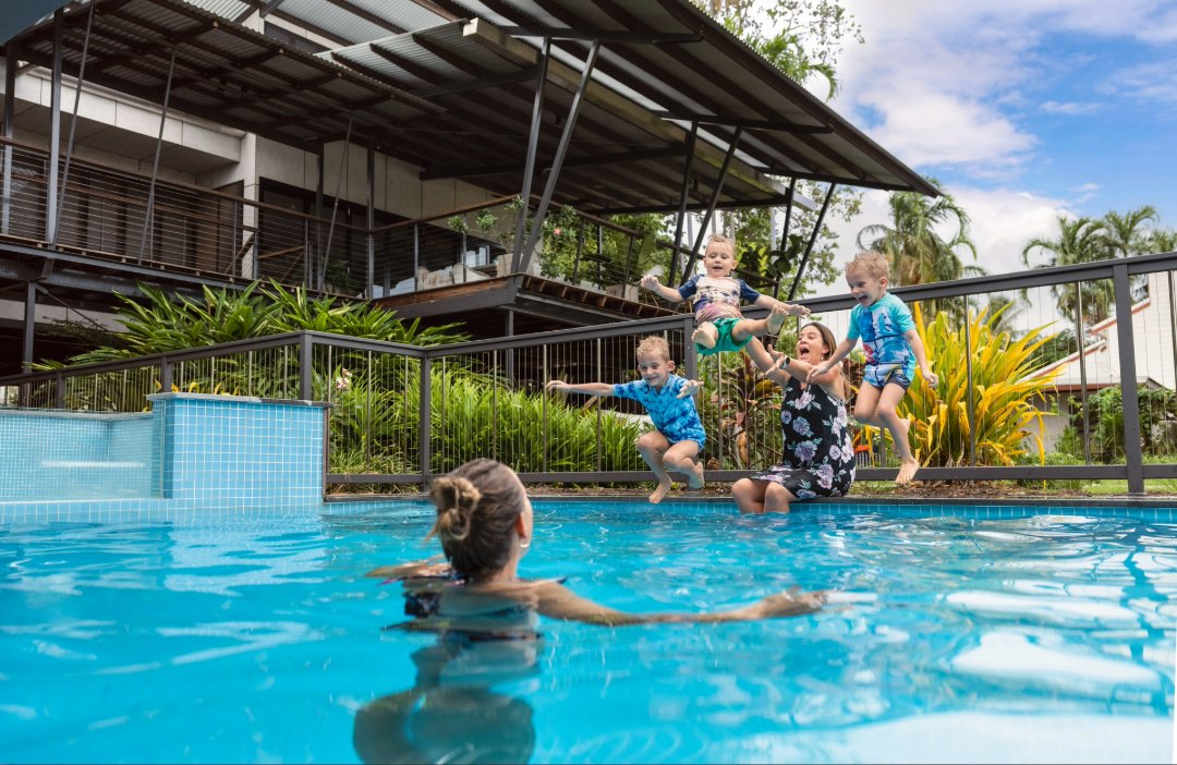 A family enjoying a sunny day by a swimming pool, with three children jumping into the water while an adult stands behind them