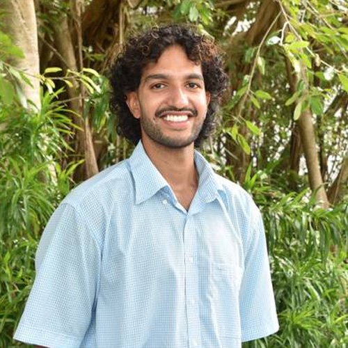 A person smiling outdoors, standing in front of lush green trees.