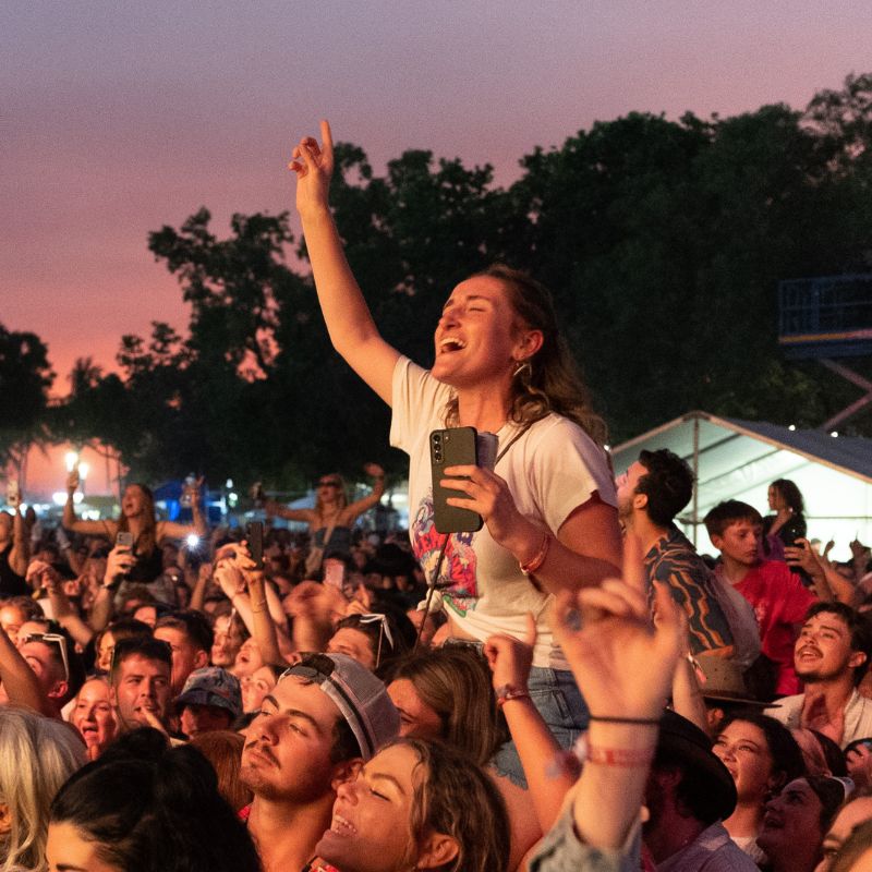 A woman sitting on someone’s shoulders cheers with her phone in hand at a crowded outdoor music event at sunset.