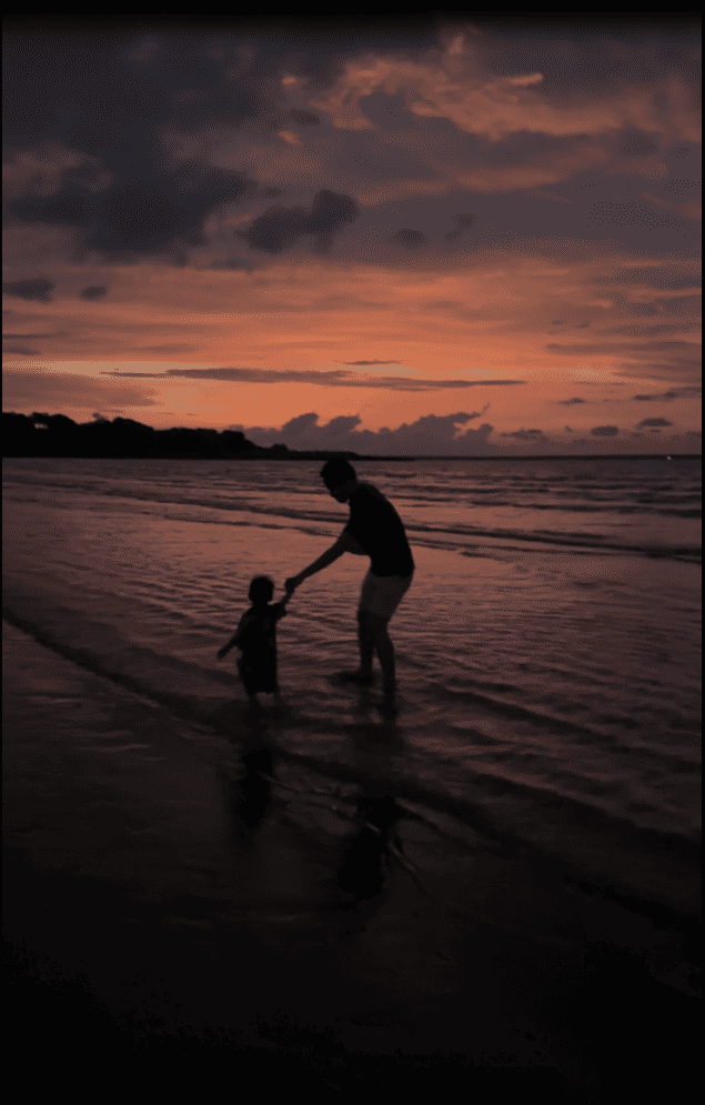 Silhouettes of an adult and child holding hands as they walk through shallow water on a beach at sunset, beneath a pink and orange sky
