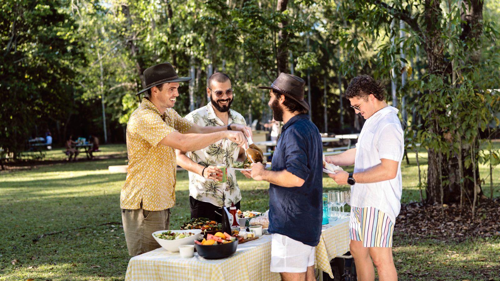 Four friends sharing food and drinks at a picnic table in a leafy park