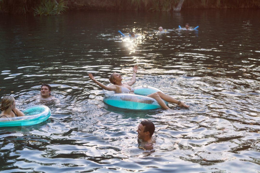 People swim in a river while others relax on inflatable tubes, with sunlight reflecting across the water.