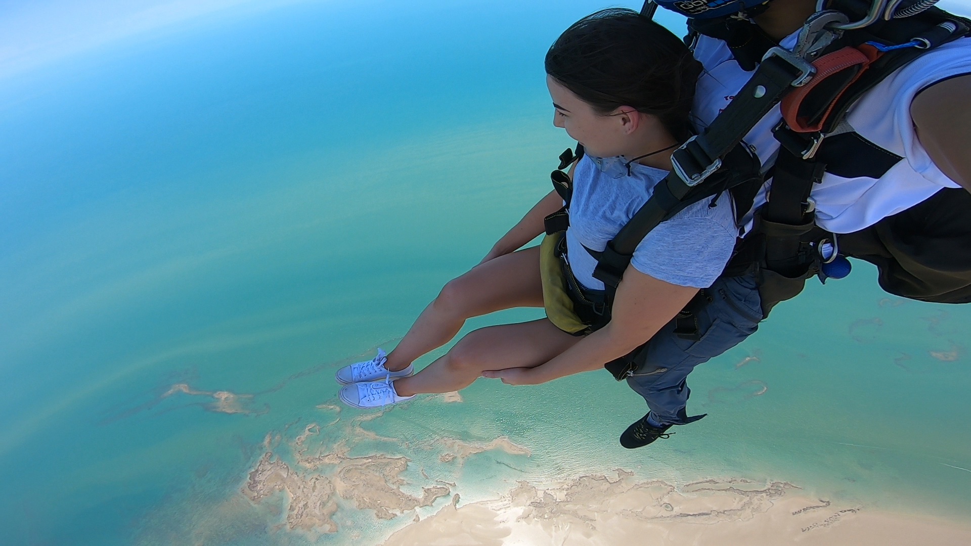Two people skydiving high above blue water and sandy islands.