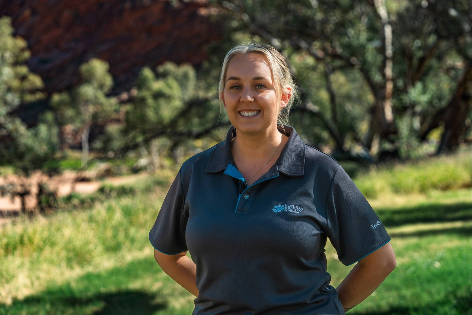 A smiling woman, Lukieda Turner, in a Northern Territory Government polo shirt stands outdoors in a sunny park.