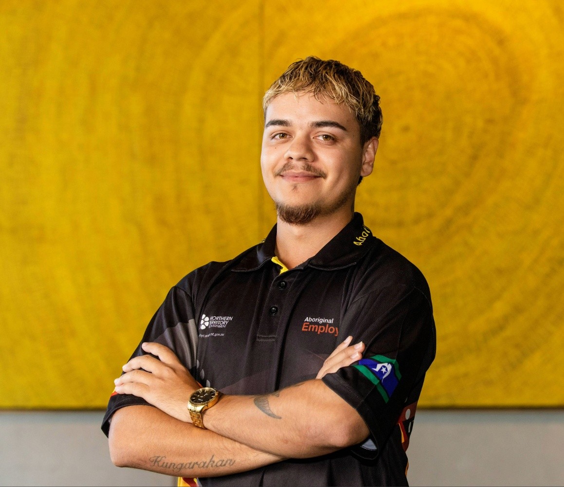 Person standing confidently with arms crossed in front of a large yellow artwork, highlighting employment and career opportunities in the Northern Territory.