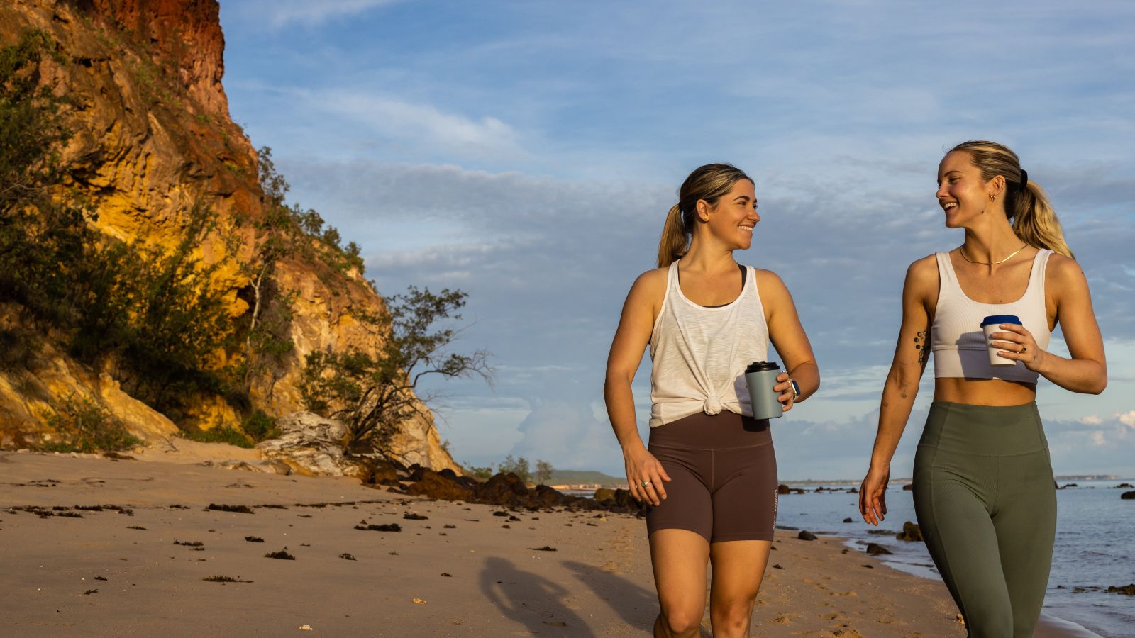 Two people in activewear holding takeaway coffees on a beach.