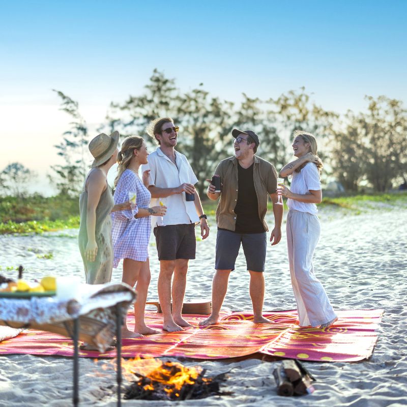 Group chatting on a beach picnic rug near a small campfire