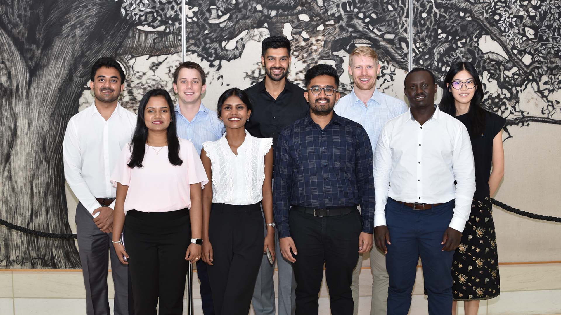 A group of eight smiling people posing in front of a tree mural.