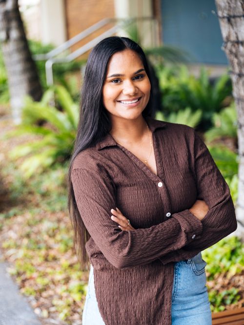 Smiling woman with long hair, brown shirt, and jeans outside.